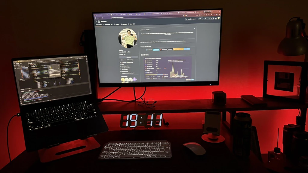 Yasin’s desk in red light, with GitHub open on the monitor, code on a laptop and a glowing digital clock